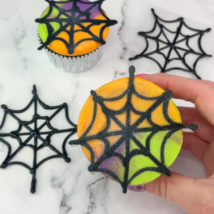 cupcake with orange, green, and purple buttercream topped with a black candy melt web and shimmer