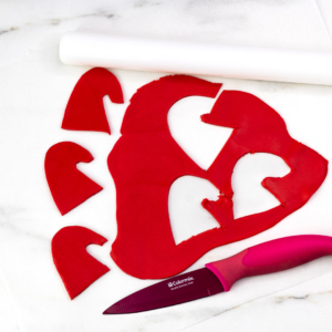 Red fondant Santa hat shapes cut out on parchment with a knife and rolling pin.