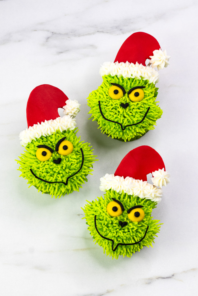 Three decorated Grinch cupcakes wearing Santa hats on a marble background. portrait