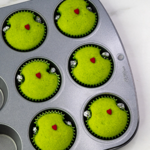 Freshly baked green Grinch-shaped cupcake with a small red heart sprinkle in the center.
