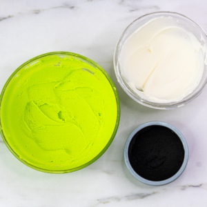 Bowls of bright green, white, and black buttercream ready for decorating Grinch cupcakes.