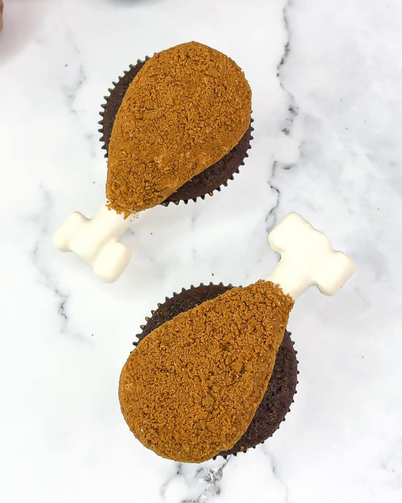 Close-up of turkey cupcakes that look like mini drumsticks, coated in crushed Biscoff crumbs with candy melt bones inserted on top. 2