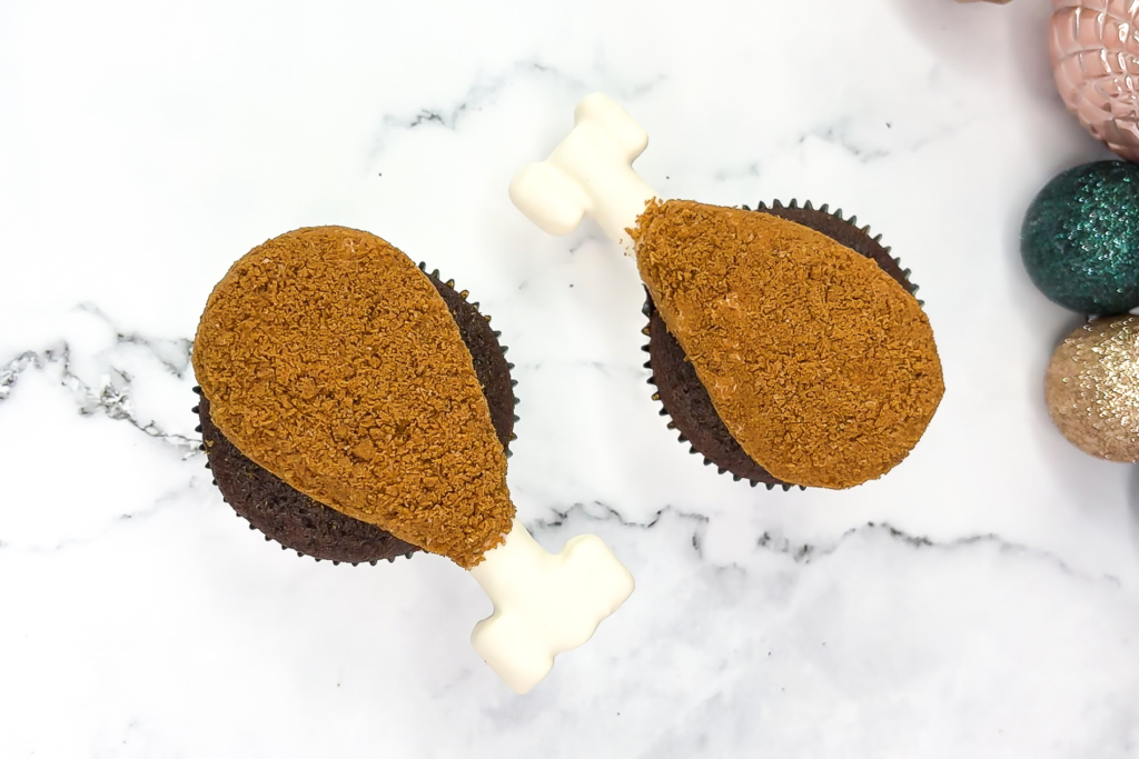 Close-up of turkey cupcakes that look like mini drumsticks, coated in crushed Biscoff crumbs with candy melt bones inserted on top. Horizontal preview