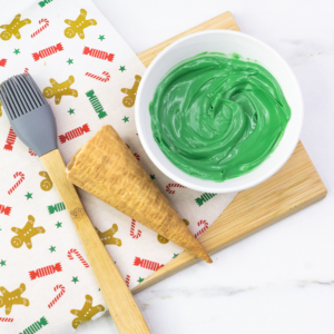 Plain sugar cone next to a bowl of melted green candy melts and a pastry brush, ready for coating.