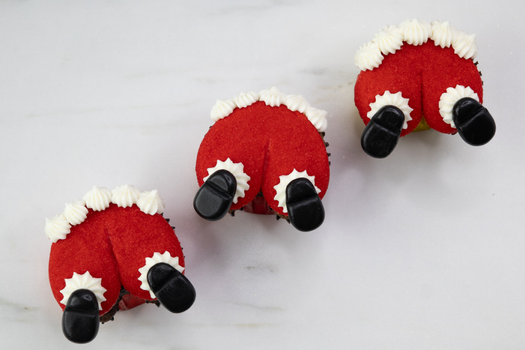 Overhead view of three Santa butt cupcakes, each coated in red sanding sugar with piped white buttercream trim and black fondant boots, arranged on a white marble surface.”