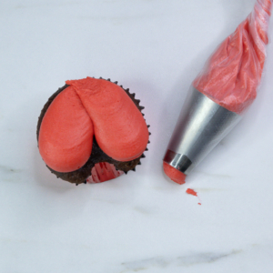 Cupcake topped with two teardrop-shaped swirls of red buttercream to form Santa’s pants, piping bag beside it.