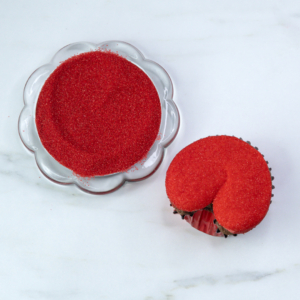 Red sanding sugar–coated Santa pants cupcake sitting next to a dish of red sanding sugar.