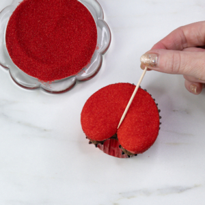 Hand using a toothpick to carve the crease line down the centre of the red sugar-coated cupcake.