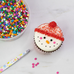 Adding pink edible confetti cheeks with tweezers to finish the snowman cupcake’s face