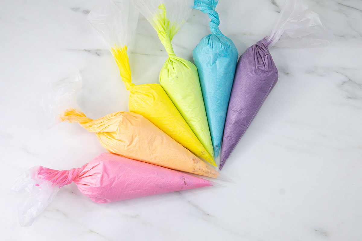 Five pastel-coloured piping bags filled with buttercream arranged in a fan shape on a marble background.